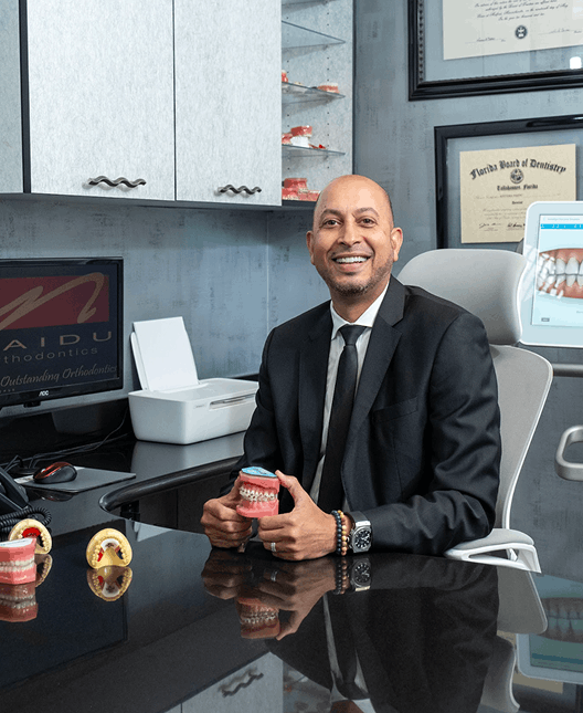A photo of Dr Naidu in office, smiling and holding dentures in his hand