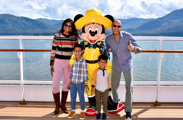 A family poses with a cheerful Mickey Mouse character on a cruise ship, with scenic mountains and water in the background.